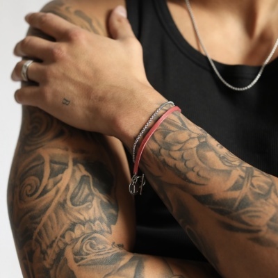 Braços tatuados de um homem com pulseiras vermelha e metálica, anel prateado e colar, veste camisola preta sem mangas