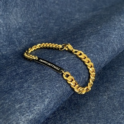 Pulseira dourada com placa preta 'TRAPPO DETTAGLI' sobre tecido azul escuro