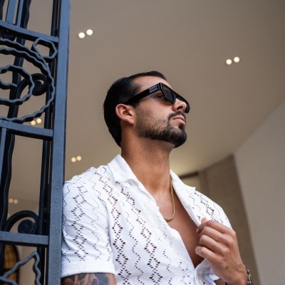 Homem com camisa branca vazada e óculos de sol pretos, apoiado em portão de ferro