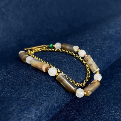 Pulseira dourada com contas cilíndricas castanhas e esferas brancas sobre tecido azul.