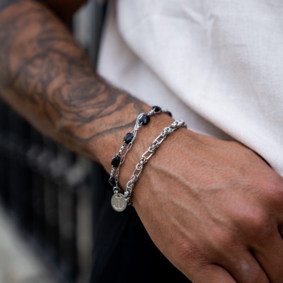 Pulseiras masculinas prateadas com contas pretas e elos entrelaçados no pulso de uma pessoa tatuada