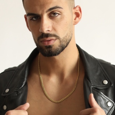 Homem com jaqueta de couro preta, colar e anel dourados, tatuagem no peito, fundo branco.