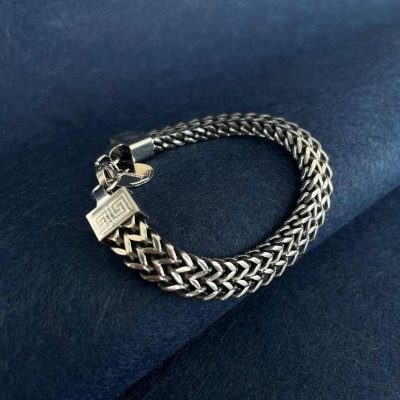 Pulseira de malha prateada com fecho retangular estampado sobre fundo azul escuro.