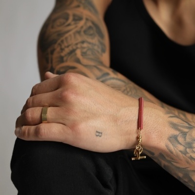 Braçadeira vermelha com fecho dourado e anel dourado na mão esquerda de pessoa com tatuagens.
