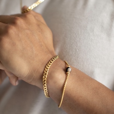 Pulseira dourada com pendente branco e corrente dourada em pulso de homem com camisa branca