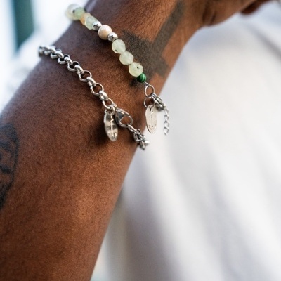 Dois pulseiras, uma de pedras verdes claras e outra de corrente prateada, num braço castanho.