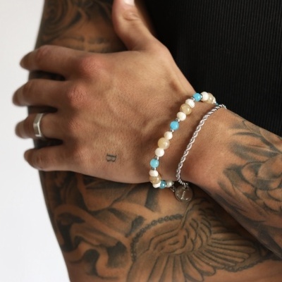 Braçadeira com contas bege e azul e pulseira prateada no braço de homem tatuado