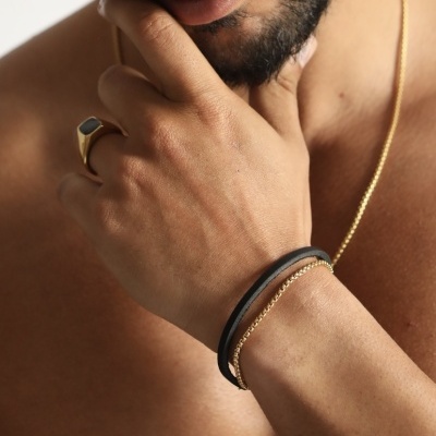 Homem com anel dourado, pulseiras preta e dourada e corrente dourada num fundo branco