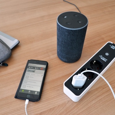 Coluna inteligente cilíndrica cinzenta, smartphone preto e régua de tomadas branca sobre mesa de madeira.