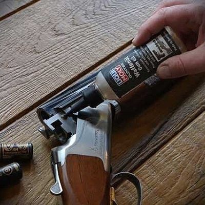 Frasco de óleo para armas HOT OIL WARRIOR junto a espingarda e cartuchos, sobre mesa de madeira.