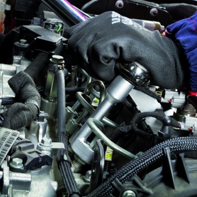 Mão com luva preta a usar ferramenta metálica num motor com cabos e tubos.