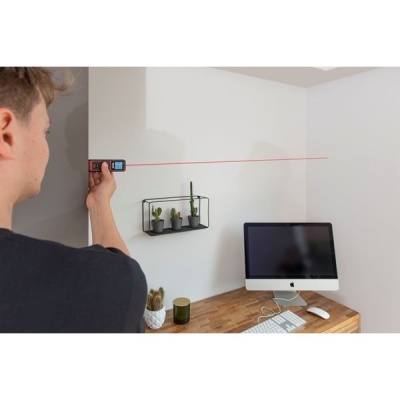 Medidor laser sendo usado para medir parede branca numa secretária com computador e plantas