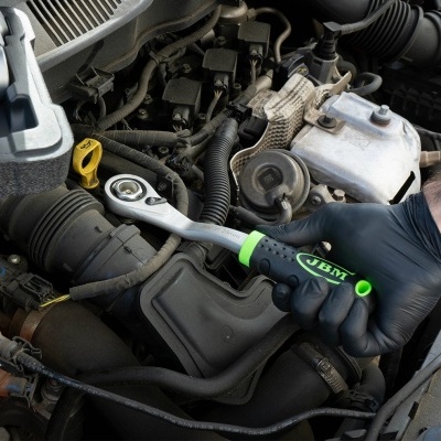 Chave de catraca JBM cinzenta e verde numa mão com luva preta a mexer num motor de carro