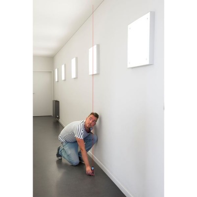 Homem mede altura da parede com ferramenta e linha laser num corredor branco.
