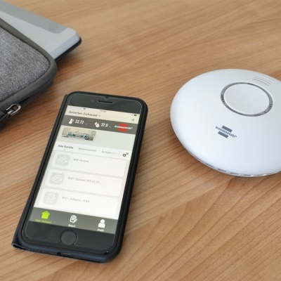 Smartphone preto com aplicação ligada e dispositivo branco redondo sobre mesa de madeira clara