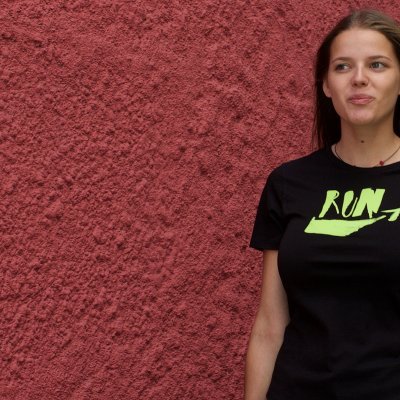 Mulher a usar uma camiseta preta com o texto RUN e setas verdes em frente a uma parede vermelho-escura
