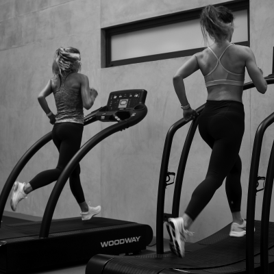 Duas mulheres a correr em passadeiras rolantes negras WOODWAY numa sala interior.