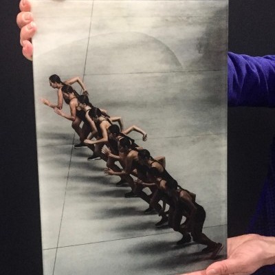 Tela com pintura de sete mulheres em posição de corrida, em tons cinza e preto, segurada por pessoa com camisola azul.