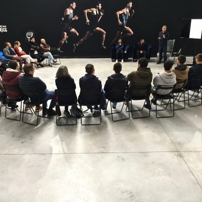 Sala ampla com pessoas sentadas em círculo e mural de corredores em fundo preto