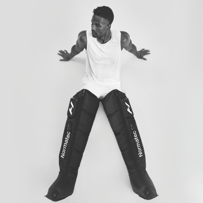 Pessoa com equipamento Normatec para recuperação nas pernas sentado contra parede branca