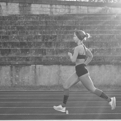 Mulher a correr numa pista de atletismo com roupa desportiva preta e ténis brancos.