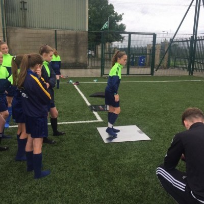 Criança em equipamento desportivo amarelo-fluorescente e azul num campo de relvado sintético com outras crianças ao redor.