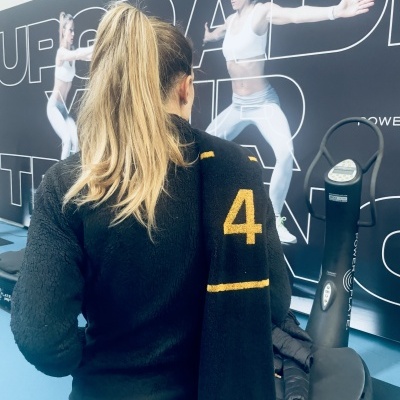 Mulher de costas em ginásio com toalha preta e painel de exercício ao fundo