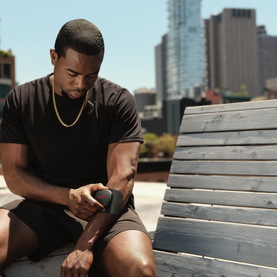 Homem sentado num banco de madeira cinzenta, vestindo roupa preta, ajustando uma pulseira com ecrã retangular e botão vermelho no pulso.