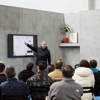 Homem a apresentar num ecrã plano para grupo sentado numa sala moderna com parede cinzenta e planta em vaso laranja.
