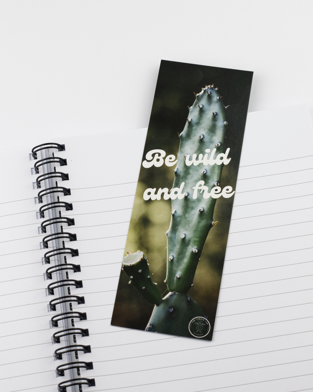 Marcador de página com cacto e texto 'Be wild and free' sobre caderno de anotações branco com linhas pretas e capa espiral preta