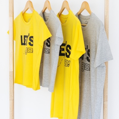 Quatro t-shirts amarelas e cinza penduradas num cabideiro de madeira com a frase LET'S GO! estampada.