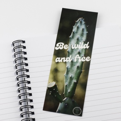 Marcador de página com cacto e texto 'Be wild and free' sobre caderno de anotações branco com linhas pretas e capa espiral preta