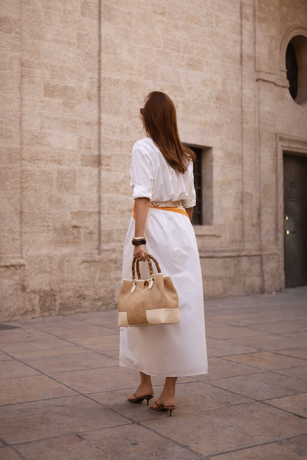 MALA EM PELE ITALIANA AMALFI RÁFIA CREAM | TUSCAN Mulher de vestido branco com acessório em ráfia e madeira, em ambiente urbano de pedra.
