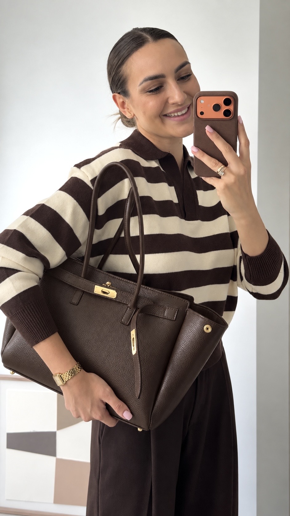MALA EM PELE ITALIANA PADOVA CHOCOLATE | TUSCAN Mulher com mala castanha de pele e camisola às riscas castanhas e creme a tirar selfie com telemóvel.