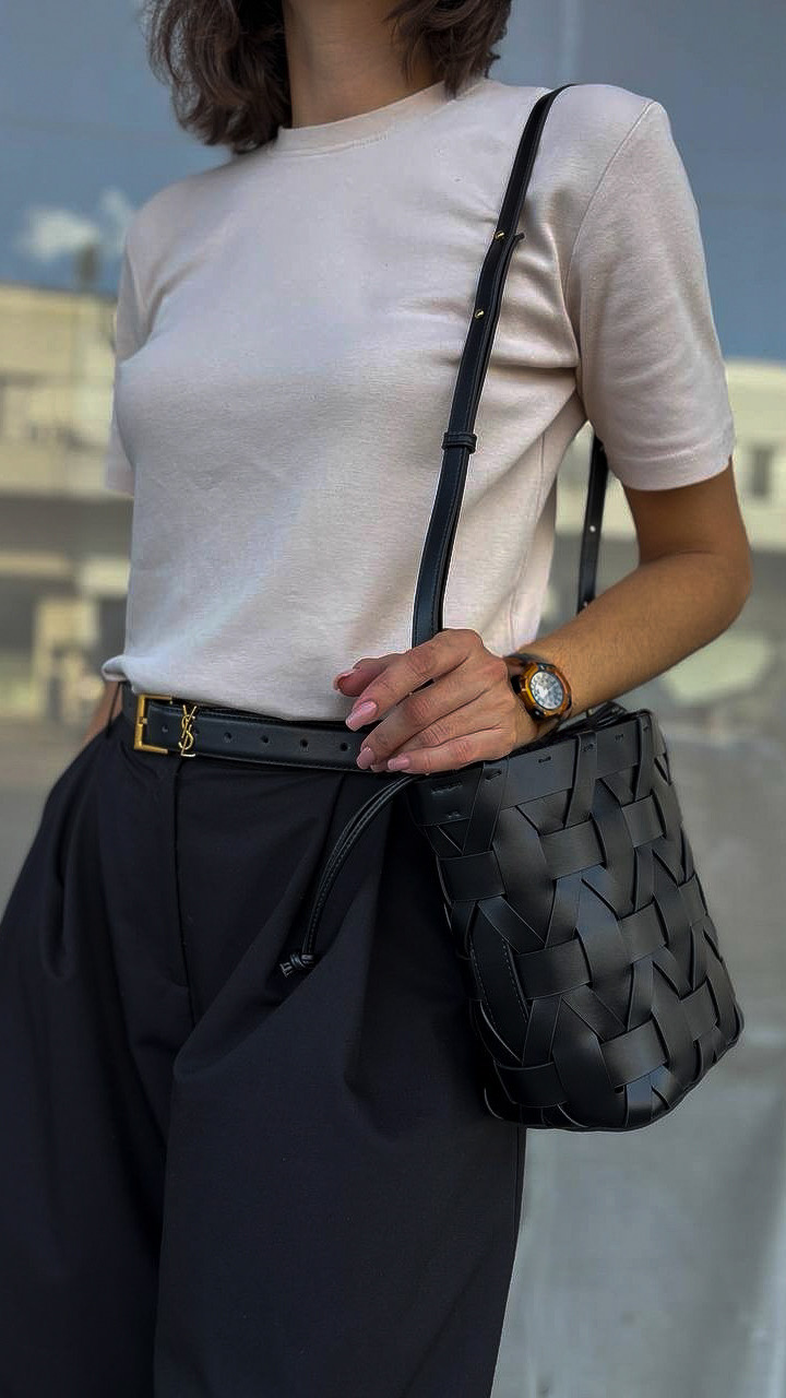 MALA DE PELE ITALIANA ENTRELAÇADA BERGAMO PRETO | TUSCAN Pessoa com camiseta branca, calças pretas e acessórios pretos incluindo cinto e bolsa entrelaçada de tiracolo