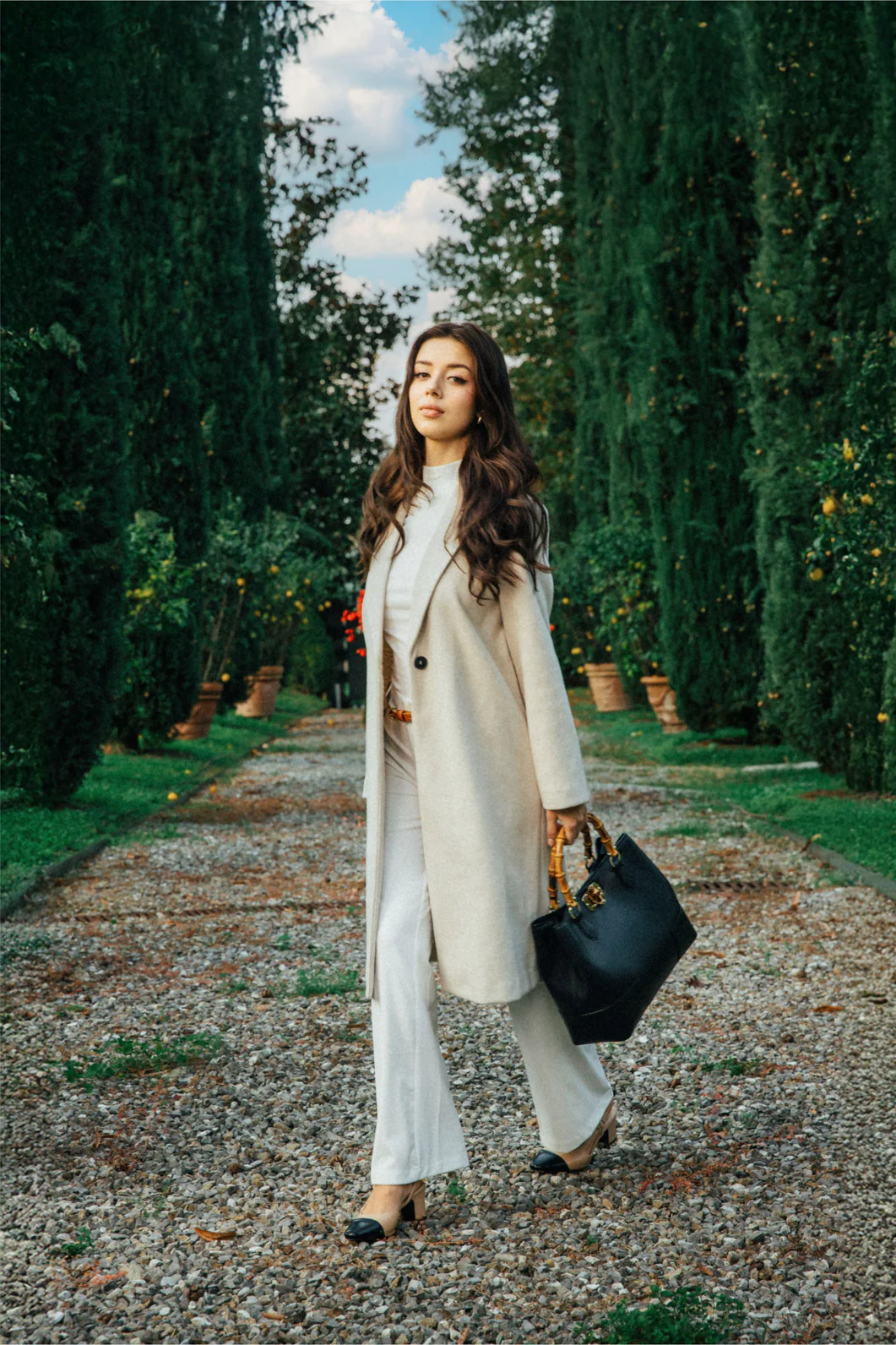 MALA EM PELE ITALIANA AMALFI PRETO | TUSCAN Mulher com casaco bege e mala preta em caminho de cascalho com árvores altas ao redor