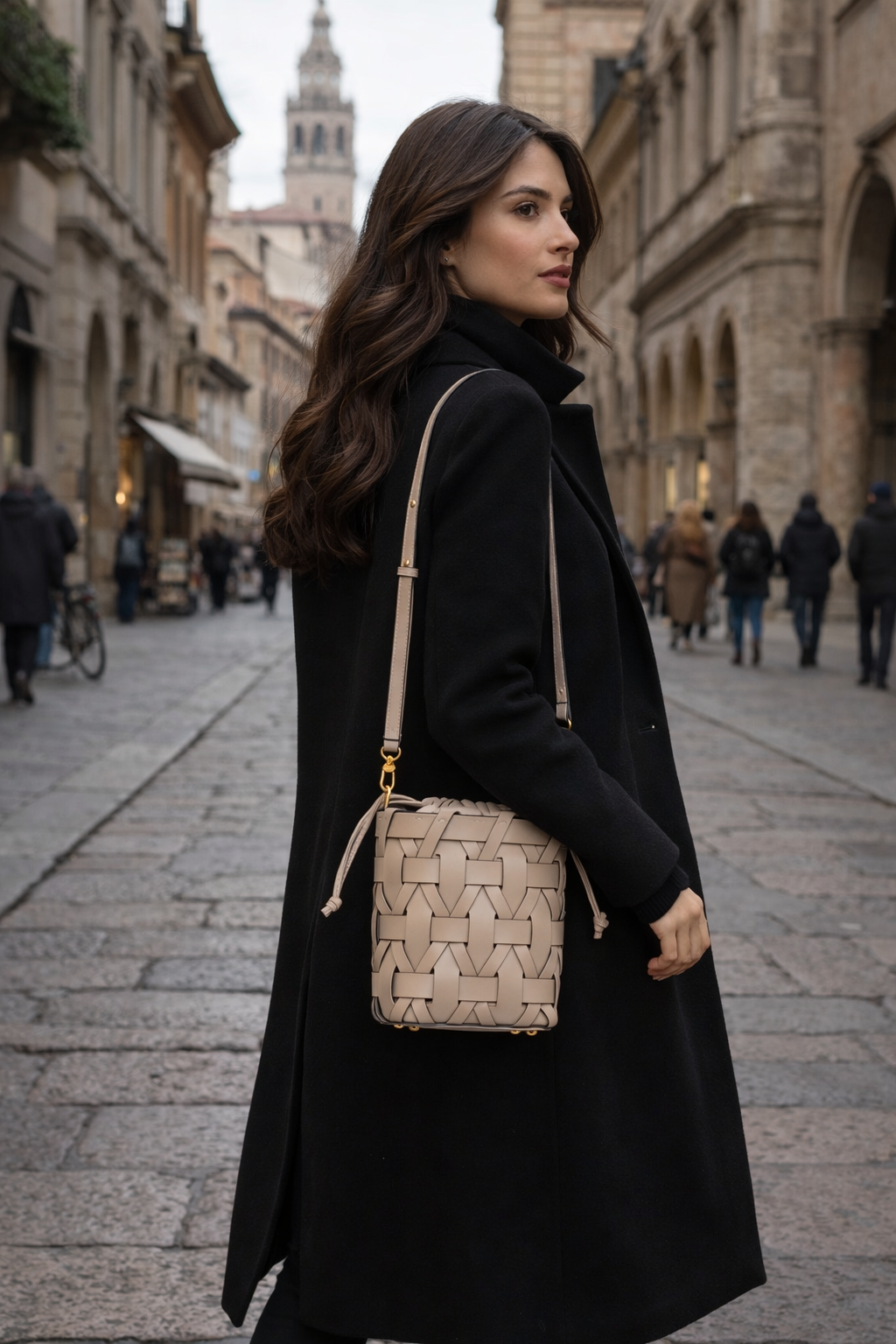 MALA DE PELE ITALIANA ENTRELAÇADA BERGAMO TAUPE | TUSCAN Mala de ombro bege com padrão trançado e alça ajustável, mulher com casaco preto numa rua de cidade histórica