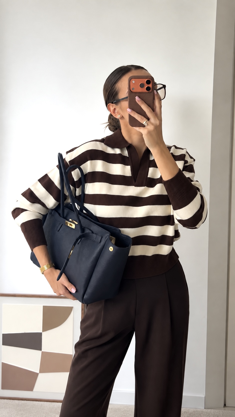 MALA EM PELE ITALIANA PADOVA AZUL NAVY | TUSCAN Pessoa vestindo camisola às riscas castanhas e creme, calças castanhas, segurando mala azul escura de couro e telemóvel com capa castanha em fundo branco.