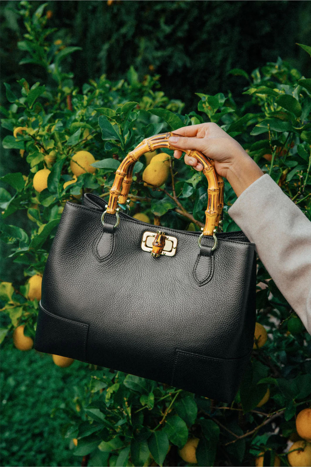 MALA EM PELE ITALIANA AMALFI PRETO | TUSCAN Mala preta de pele com pegas em bambu e fecho dourado em frente a limoeiros