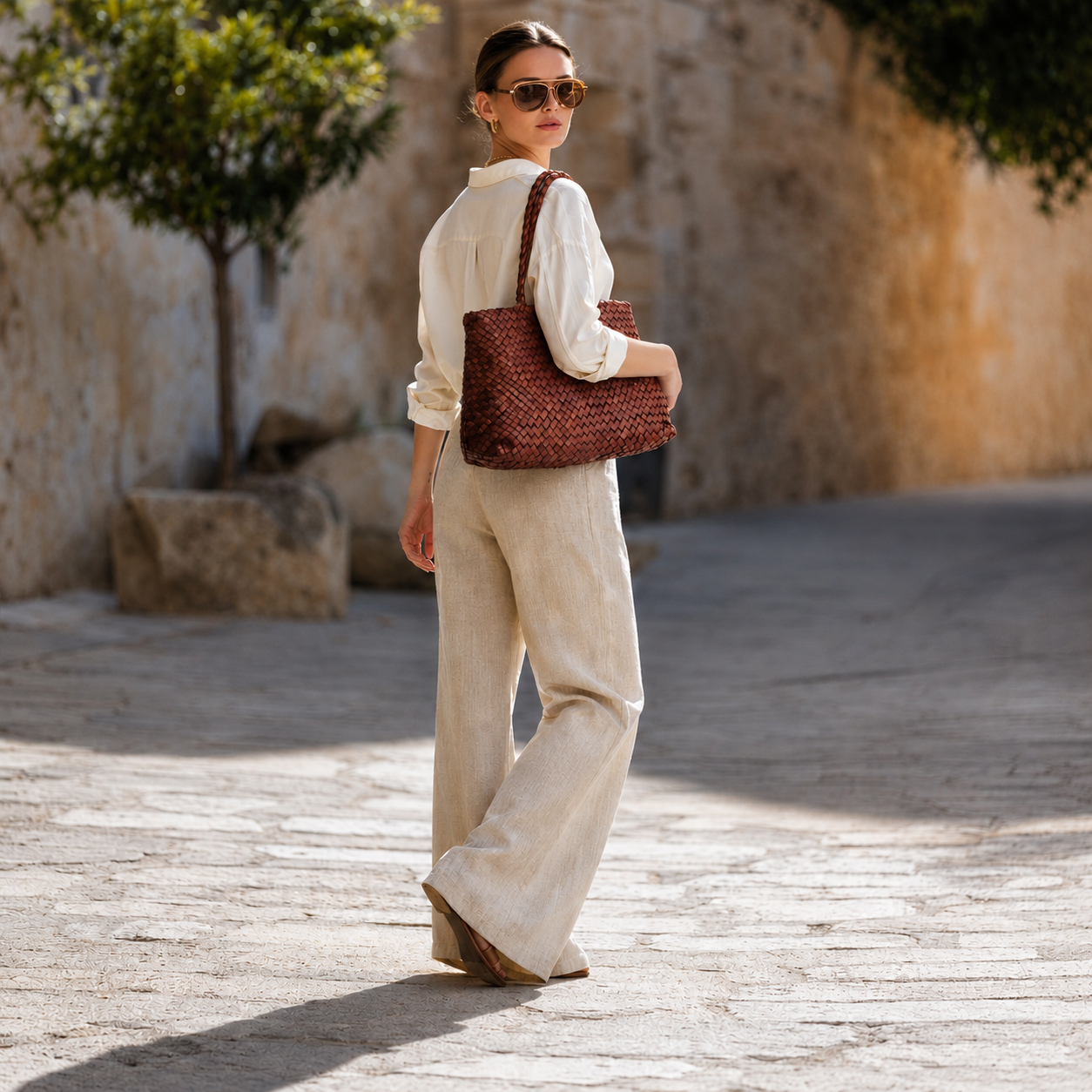 MALA DE PELE INTERCCIATO ITALIANA SORRENTO | EDIÇÃO EXCLUSIVA Mulher com camisa branca e calças largas bege, usando mala de ombro castanha texturizada e óculos de sol castanhos, em rua com parede de pedra.