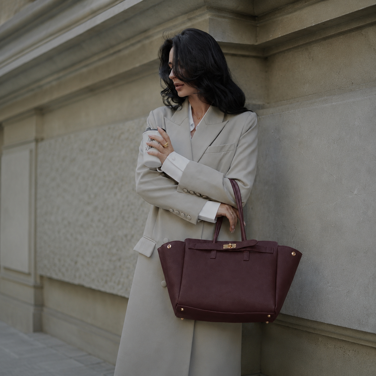MALA EM PELE ITALIANA PADOVA BURGUNDY | TUSCAN Mulher com casaco bege claro e saco castanho-escuro junto a parede de pedra cinza.