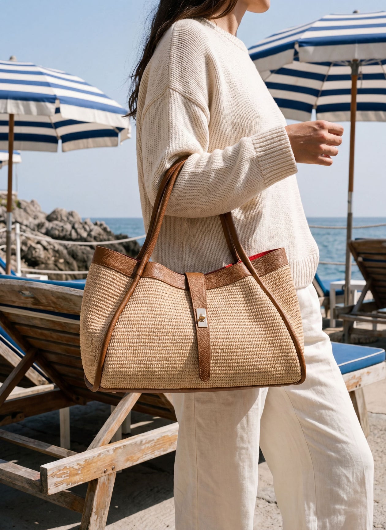 MALA EM PELE & RÁFIA ITALIANA ROMA | TUSCAN Bolsa de ráfia e pele castanha com fecho metálico dourado, usada com camisola creme e calças claras, em ambiente de praia com espreguiçadeiras e chapéus de sol às riscas azuis e brancas
