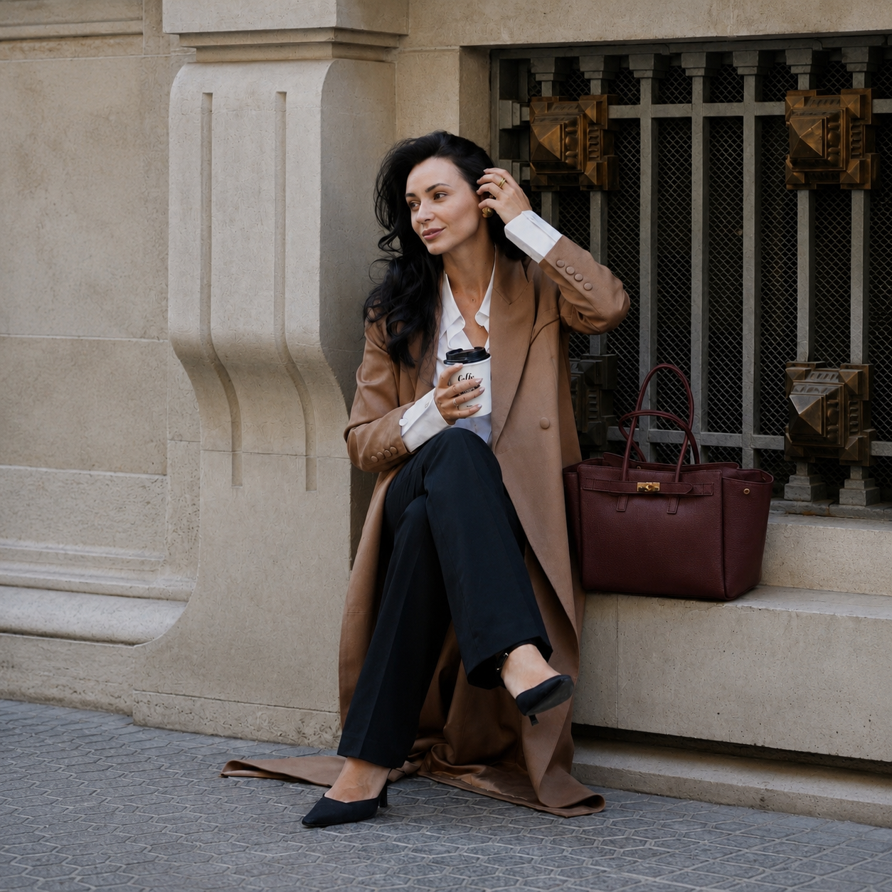 MALA EM PELE ITALIANA PADOVA BURGUNDY | TUSCAN Mulher sentada na calçada com casaco camel, calças pretas e mala castanha