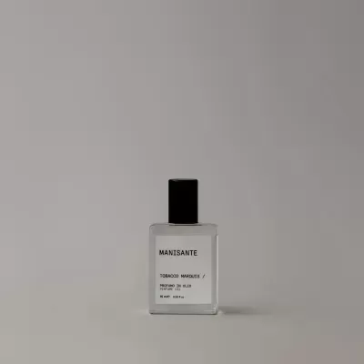 Frasco de perfume MANISANTE transparente com rótulo branco e tampa preta sobre superfície cinzenta.