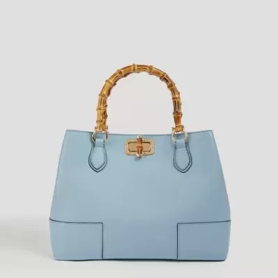 Mala azul claro em pele com alças de bambu e fecho dourado sobre fundo cinza claro