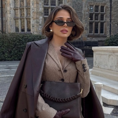 Mulher estilosa com casaco castanho, fato xadrez, luvas, bolsa de pele castanha e óculos de sol pretos em cenário urbano