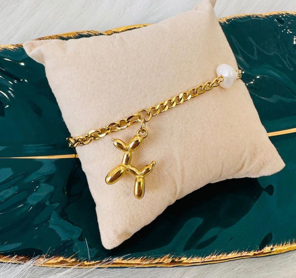 Pulseira dourada com pendente em formato de balão de animal sobre almofada bege