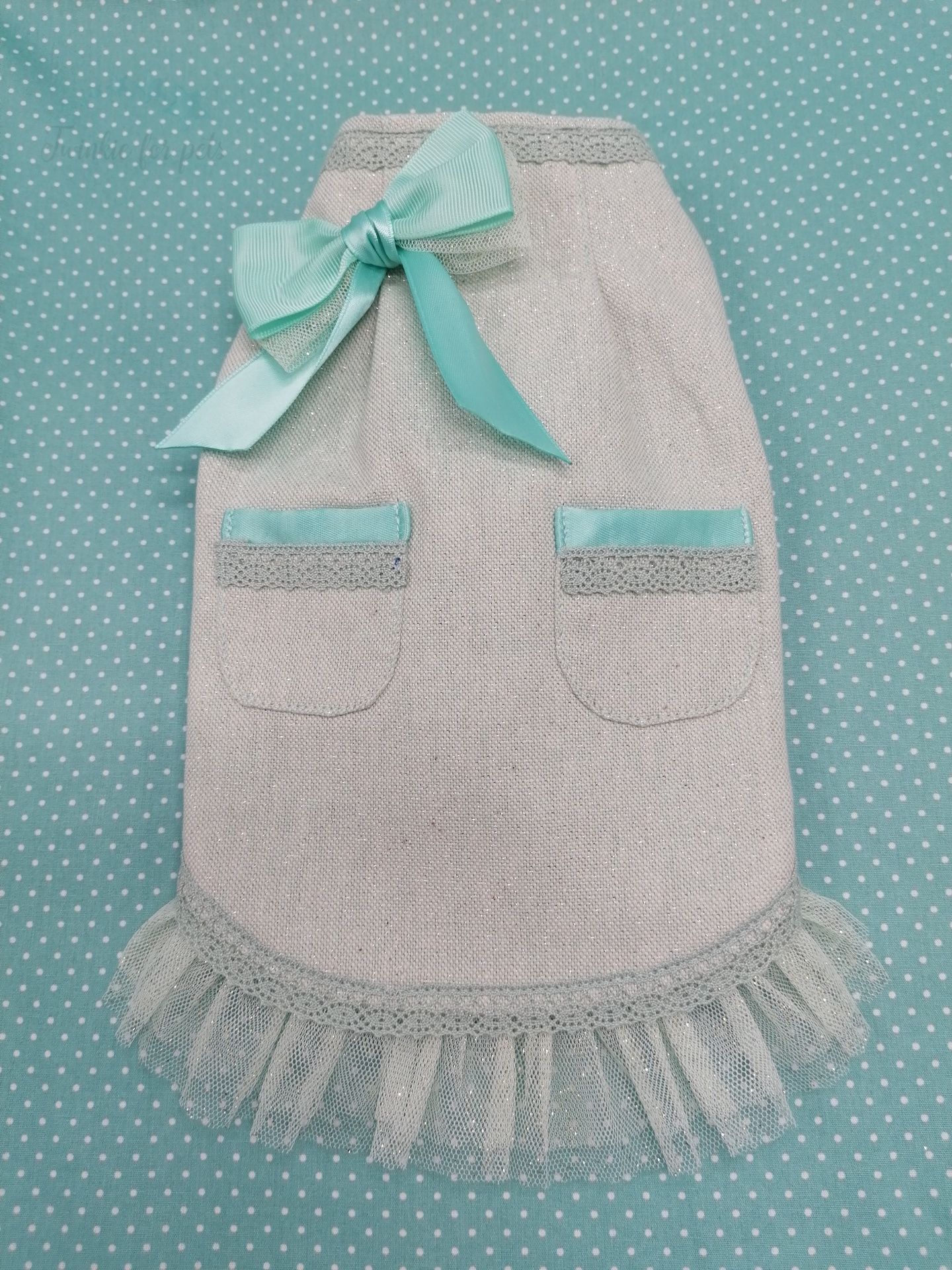 Roupa para cão bege com bolsos e fita azul clara sobre tecido azul com pontos brancos.