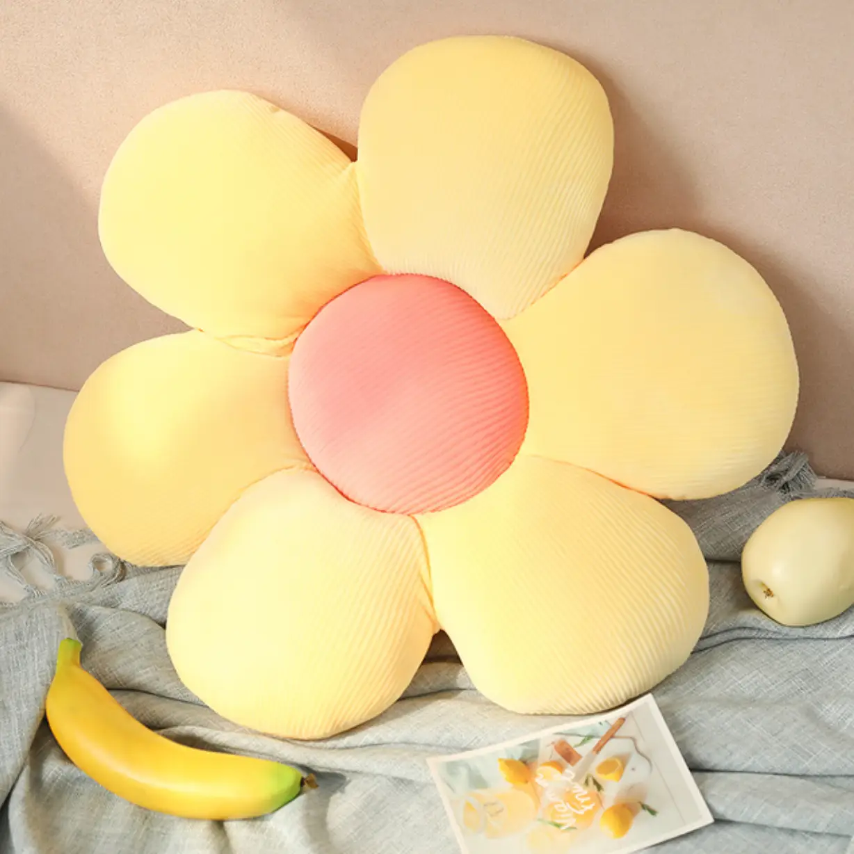 Almofada em forma de flor amarela com centro rosa, sobre tecido cinzento, com uma banana e uma maçã ao lado