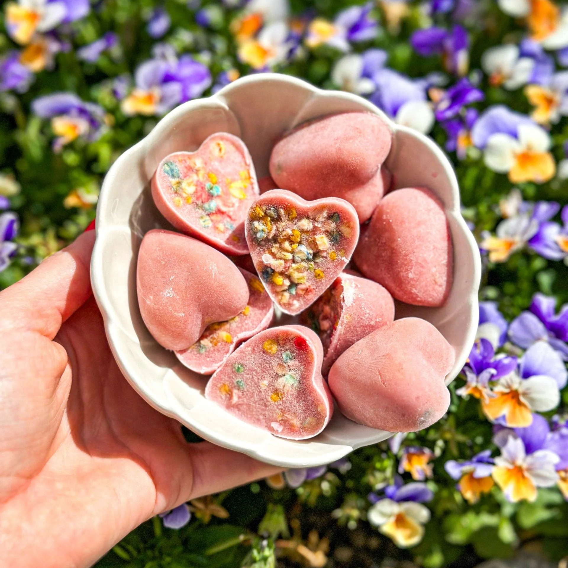 Taça branca com corações rosa decorados, mão a segurá-la, flores coloridas ao fundo